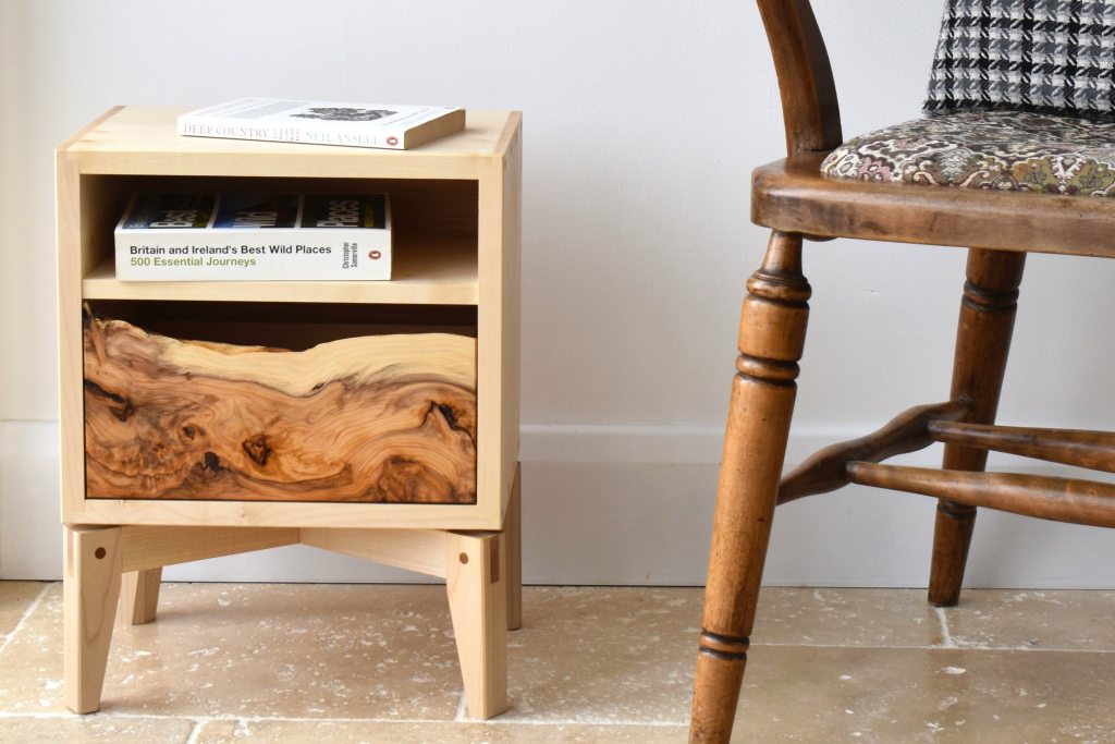 A wooden nightstand with a natural wood drawer and an open shelf, displaying two books on top, next to a vintage wooden chair.