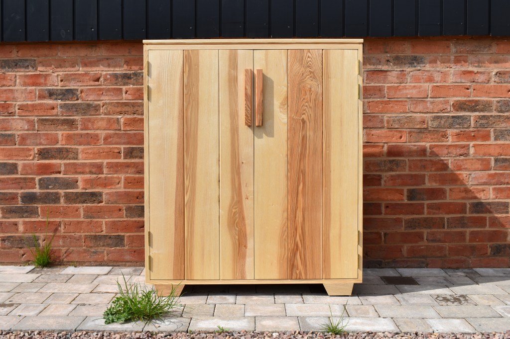 A wooden cabinet with a natural finish, featuring two doors and a rectangular shape, placed against a brick wall.